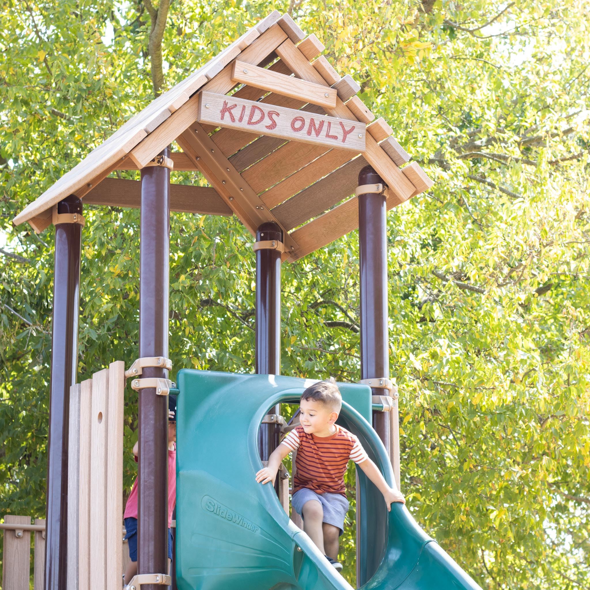 Boy on top of a slide looking down smiling; the sign over his head reads "KIDS ONLY"